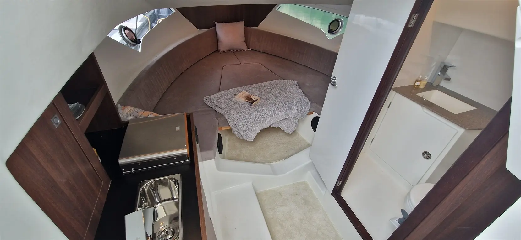 Interior view of a modern boat cabin with a circular seating area, kitchenette, and bathroom, featuring a blanket and book.